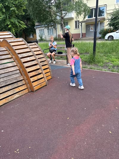 The image depicts two children and a young woman outdoors on a playground. The scene is set in Verkhnerussky, Russia, with apartment buildings visible in the background. The ground is covered in a dark reddish-brown material, likely a rubberized surface common in playgrounds. To the left, a curved wooden structure with multiple planks creates a stepped area.

In the foreground, a young girl wearing blue overalls over a pink shirt and white sneakers is standing on the rubberized surface, facing away from the camera. Her blonde hair is tied in a braid. To her left, a young woman wearing a black t-shirt, brown pants, and a light green cap is sitting on a wooden bench, looking towards the girl and gesturing with her left hand. She has long blonde hair. Slightly further back and to the left, another child, a boy with blonde hair, is sitting on the same bench. He is wearing a white and blue striped t-shirt with black shorts and white sneakers, and he is holding a dark object that resembles a phone.

The lighting suggests it is daytime, likely overcast given the absence of strong shadows. The presence of green grass and trees indicates a warm season. The overall mood seems relaxed and casual, suggesting a typical day at the playground. No visible text is present in the image.