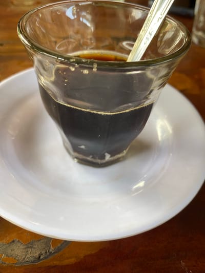 A close-up, overhead shot shows a clear glass of dark coffee with a small spoon inside. The glass is resting on a white saucer, which is on a wooden table. The coffee has a frothy layer on top, and small bubbles are visible on the inside of the glass. The spoon is silver and has a decorative pattern on its handle. The wooden table has a textured surface with variations in color, suggesting it is aged or made of a natural wood. The lighting in the image is soft, with reflections visible on the glass and saucer. The image appears to be taken in Aluepiet, Indonesia, likely in a cafe or dining setting.