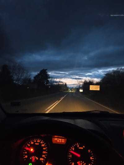 The scene depicts a view from the interior of a vehicle driving on a paved road at dusk or early night in Novi Chiflik, Bulgaria. The sky is largely dark and overcast with prominent gray clouds, transitioning to lighter blue and faint orange hues low on the horizon. The road ahead is illuminated by the vehicle's headlights, revealing double solid white lines marking the center of the two-lane asphalt surface. Dark, silhouetted trees line both sides of the road, with some distant lights, likely streetlights or from a settlement, visible further down the road. On the right side of the road, a yellow rectangular sign with dark text is partially visible. The vehicle's dashboard is in the foreground, illuminated with red backlighting. The speedometer on the left indicates a speed of approximately 70 km/h, with a red warning light illuminated near 0 km/h and a green light at the bottom. The tachometer on the right shows the engine speed just below 2000 RPM. A digital display between the gauges reads "288 540 Km" for total mileage and "97.5 Km" for a trip odometer. The top edge of a dark steering wheel is visible at the bottom center of the frame. No individuals are visible.