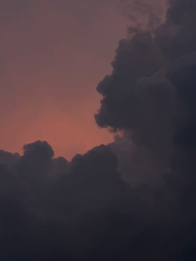 A dusky sky with hues of rose and deep indigo serves as the backdrop for a dramatic formation of dark, heavy clouds. The clouds, dense and textured, occupy the majority of the frame, obscuring much of the sky. Their varied shapes and shadows create a sense of depth and volume. The lighting suggests either early morning or late evening, with a soft, diffused glow emanating from behind the cloud mass on the left, casting a warm, reddish-pink light onto the thinner portions of the clouds and the sky itself. The overall impression is one of a powerful, perhaps impending, weather event.