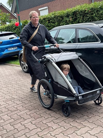 A woman with light blonde hair pulled back, wearing round glasses, a black long coat, grey pants, and black Converse-style sneakers, pushes a black and grey "Thule Chariot Sport" child carrier on a paved pathway in Deinste, Germany. A young boy with light brown hair and dark-rimmed glasses sits inside the carrier, looking forward. The woman has a light brown messenger bag slung over her right shoulder.

To the left, a blue Ford Puma SUV with German license plate "STD UN 81" is parked. To the right, a black SUV or wagon with raindrops on its windows is parked. A green pole with a red and a light blue balloon attached stands behind the woman, positioned between the two parked vehicles. Behind the cars, a dense green hedge lines the background, with a brick building featuring a dark roof and a white roll-down shutter partially visible. The ground is covered with light reddish-brown interlocking pavers. The scene suggests an overcast day.