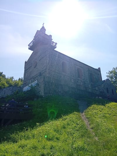 A stone church with a bell tower is situated on a grassy hill under a bright, sunny sky. The church features arched windows and a cross at the top of the bell tower. A banner is visible on the side of the church. In the foreground, several people are seated on a wooden structure. Lush green grass and vegetation cover the hill leading up to the church. A dirt path winds its way up the hill. The sun is bright, causing some lens flare in the upper part of the image. This is the Church of the Assumption of the Blessed Virgin Mary in Sobótka, Poland.
