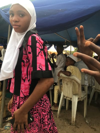 A girl in a white hijab and a pink and black patterned dress sits in front of a group of people. Some individuals are wearing white skullcaps and light-colored clothes. One person is seen making a hand gesture in the foreground. White plastic chairs are stacked nearby, and a blue tent or canopy is visible in the background. The ground appears to be sandy.