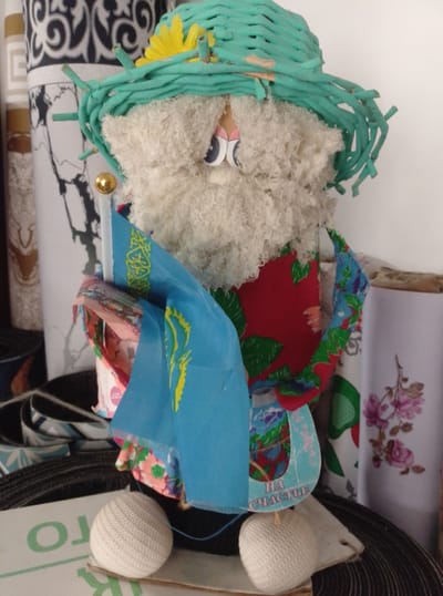 The image shows a handcrafted doll. The doll is wearing a green woven hat adorned with a yellow flower and has a shaggy white beard and large cartoon-like eyes. It is holding a blue flag with yellow symbols, possibly the flag of Kazakhstan, and a decorative element with the Cyrillic text "НА СЧАСТЬЕ" (For Happiness). The doll is dressed in a red and green patterned fabric. It is standing on a coiled black material, and in front of it are two white spherical objects that appear to be feet or decorative elements. In the background, there are rolls of fabric with patterns, including one with a floral design and another with a black and white abstract pattern. The doll is placed indoors, possibly on a shelf or table, in an environment that suggests a domestic setting or a craft display. There is no discernible connection to the city of Şemonaïxa, Kazakhstan, other than the potential national symbol on the flag.