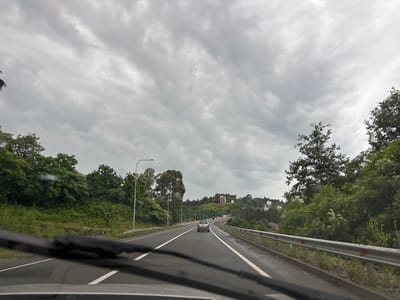 The image captures a view from inside a car, looking forward through the windshield. The car is on a multi-lane highway with a grey, overcast sky above. The highway is bordered by lush green trees and vegetation on both sides, with a metal guardrail visible on the right. In the distance, there are buildings situated on rolling hills, suggesting an urban or semi-urban environment. A silver car is driving ahead in the same lane, and other vehicles are visible further down the road. The weather appears to be cloudy, possibly with light rain, as indicated by the wet appearance of the road and the streaks on the windshield. The overall mood is somber, likely due to the cloudy weather. The time of day is unclear, but the light suggests it could be morning or afternoon. The location is identified as Makhinjauri, Georgia. There is no visible text in the image.