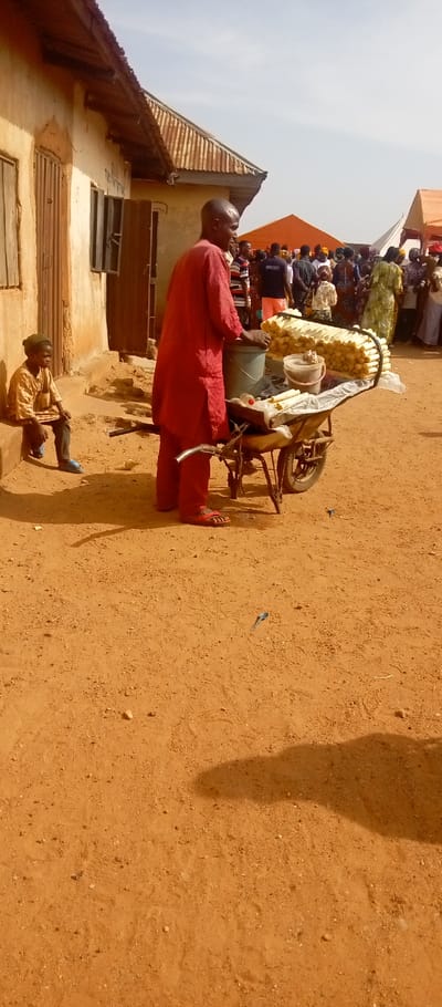 The image captures an outdoor scene in Kasuwan Magani, Nigeria, under a clear, hazy sky. The ground consists of reddish-brown dirt, with a metallic spoon-like object and a small blue item visible on its surface. In the center, a man in a red tunic and trousers, wearing red sandals, stands with his back partially to the viewer, facing a two-wheeled pushcart. The cart is laden with goods, including a grey bucket, a lidded plastic container, a white plastic bottle, and multiple rows of horizontally stacked, cylindrical, pale yellow wrapped food items. To the left, a young person dressed in a light brown shirt, dark trousers, and light blue sandals sits on a raised, light-colored step of an adjacent building. This building features a weathered wooden door and open windows with wooden grilles, topped by a corrugated metal roof. In the background, a large group of people is gathered, and several tents, including a prominent orange canopy, are visible above the crowd.
