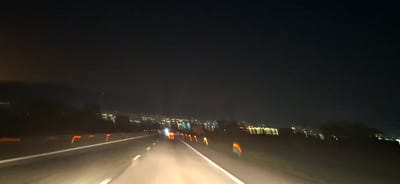 The image depicts a night view of a highway, likely within or near the city of Yana, Bulgaria, given the context. The road is illuminated by the headlights of the vehicle from which the photo was taken, creating bright streaks of light on the asphalt. The highway is marked with white lines and bordered by orange reflective markers on the right side, indicating the edge of the road. In the distance, a city skyline is visible, characterized by numerous glowing lights that suggest buildings and streetlights. The sky is dark, indicating nighttime, and there appears to be a faint haze or fog that slightly diffuses the distant lights. The overall scene is one of a vehicle traveling on a highway at night, with a city illuminated in the background. There are no people or specific actions clearly discernible in the image, only the environment of a road at night with distant urban lights.