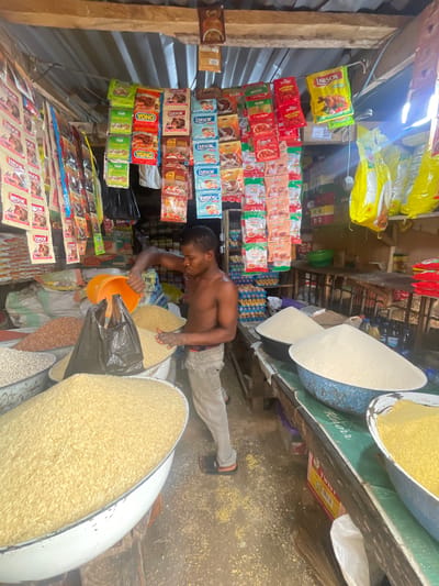 In the city of Ojodu, Nigeria, a shirtless man in beige pants fills an orange bucket with grain from a large bowl. The scene is inside a small, cluttered shop. Various packaged food items, including spices and seasonings like "Arsor," "Onga," and "Gino," are displayed on the walls. Baskets filled with different types of grains are in the foreground. The shop appears to be a local market or provision store.