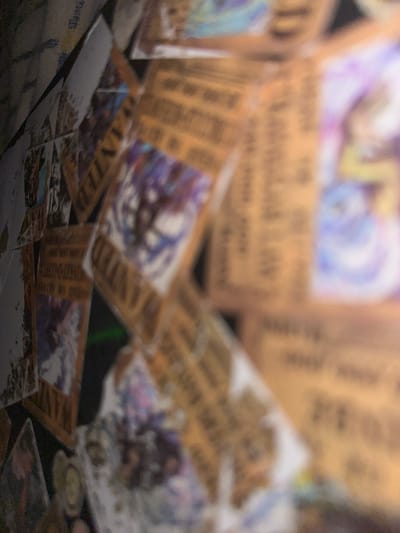 The image displays a collection of WANTED posters, some of which are partially obscured. The posters are predominantly a light brown or tan color, with text and illustrations visible on them. Some posters feature images of anime-style characters. The lighting in the image is dim, with some areas brightly illuminated and others in shadow. The overall impression is that of a display or collection of these posters. There is no indication of the location being Nizhneye Myachkovo, Russia, as no specific environmental or identifying features are present to suggest this. The scene appears to be indoors, with the posters arranged in a somewhat haphazard manner, overlapping each other. No people are visible in the image.