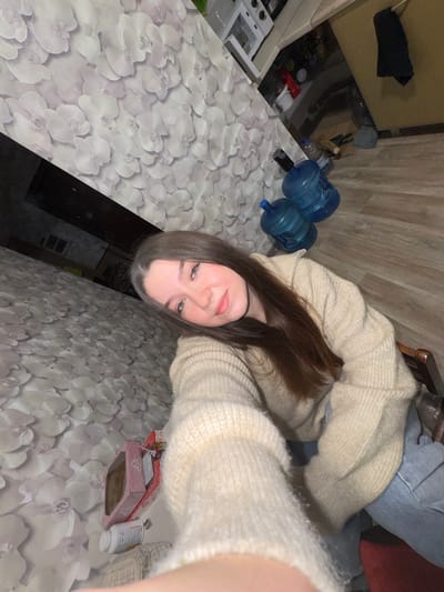 The image depicts a young woman with long, brown hair, wearing a beige knit sweater and blue jeans. She is seated indoors, with a patterned wallpaper visible on the left side of the frame. In the background, there are two large blue water bottles, a broom, and what appears to be a kitchen counter with some items on it. The flooring is wooden. The overall impression is a casual indoor setting. The location is not specified as Башкултаево, Russia.