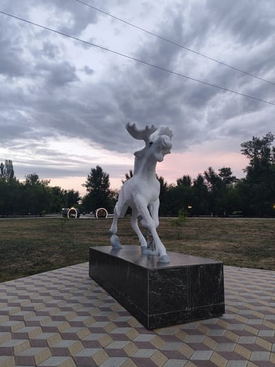 A white statue of a moose is the central subject of this image, set against a backdrop of trees and a cloudy sky at dusk. The moose sculpture is depicted in a dynamic pose, with its head tilted and antlers spread, appearing to be in motion. It stands on a dark marble pedestal, which rests on a patterned brick walkway.
In the background, a line of trees forms a dark silhouette against the twilight sky, which exhibits soft hues of pink and gray. There are illuminated circular structures visible in the middle ground, suggesting a park or recreational area. The ground is covered with dry grass, indicating it may be late summer or early autumn. The overall mood is serene and slightly melancholic, characteristic of a fading day. The time of day is dusk, with overcast weather contributing to the subdued lighting. No visible text is present in the image. The location context is Slavgorod, Russia.