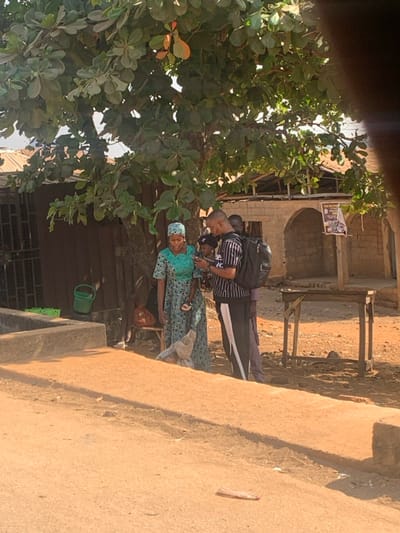 The scene depicts a group of four individuals standing under a large deciduous tree on an unpaved, reddish-brown dirt ground in Egbedi, Nigeria, during daylight hours.
In the center-left, a woman in a teal-green patterned long dress and matching headwrap holds a small, dark-skinned child in her left arm. She is smiling and looking towards a man to her right. The child, wearing dark clothing and light sandals, is also looking towards a mobile phone.
To the right of the woman and child, a man with a shaved head wears a black and white striped short-sleeved shirt, dark pants with a white side stripe, and a black backpack. He is holding a black mobile phone, presenting its screen to the woman and child. Partially obscured behind this man is another man in a dark shirt.
The group appears to be engaged in viewing content on the mobile phone.
In the left background, a dark, corrugated structure with a barred window is visible. A green plastic bucket and two green crates are positioned near this structure. In the right background, a light-brown, partially dilapidated brick building with two arched openings and exposed wooden roof beams stands. A wooden pole with a poster is affixed in front of this building.
A concrete curb or low wall runs across the midground from left to center. A simple, four-legged wooden table is situated in the right midground. A small, light-colored piece of debris lies on the dirt ground in the foreground.