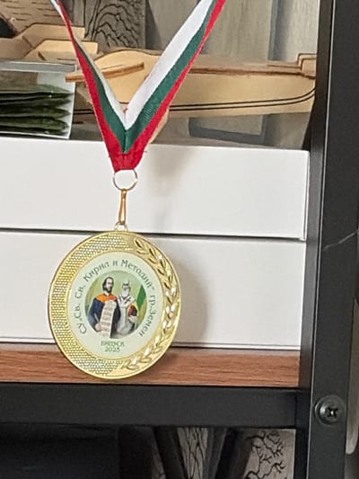 A close-up, eye-level shot shows a gold-colored medal hanging from a tricolor ribbon, superimposed on a shelf with white boxes. The medal, circular with a textured gold border, features an image of two figures, likely saints, in traditional attire. Text in Cyrillic encircles the image, reading "Св.Св. Кирил и Методий" (Ss. Cyril and Methodius) and "гр.Земел" (town Zemel). Below the figures, the year "2025" is visible. The ribbon, composed of alternating red, white, and green stripes, hangs vertically. In the background, a partial view of a wooden model or decoration is visible, with stacked green items in a transparent container to the left. The medal and ribbon are the primary focus, positioned slightly right of center.