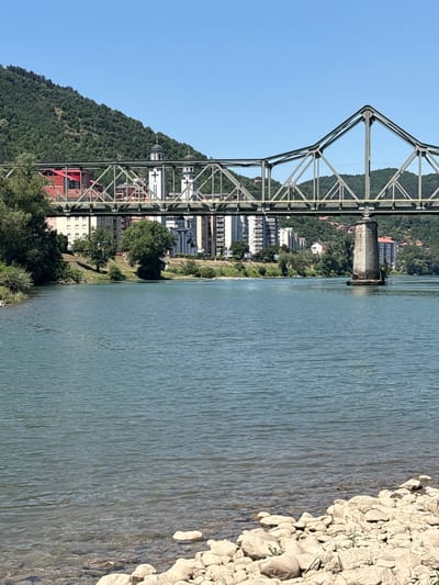 An eye-level, medium shot captures a bright, sunny day at the Drina River in Zvornik, Bosnia and Herzegovina. A multi-span steel truss bridge, painted in a muted green, dominates the upper portion of the frame, spanning the wide, calm blue river. On the far bank, a densely wooded, green hillside rises steeply under a clear, light blue sky. A line of buildings, including modern high-rises and what appears to be a church with a white steeple and domes, are visible nestled at the base of the hill, behind the bridge. The water surface is rippled, and a rocky, light-colored shoreline occupies the bottom of the image.