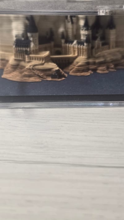 The image shows a layered model of a castle, possibly Hogwarts from Harry Potter, enclosed in a clear plastic case. The castle model is made of what appears to be cardboard or paper, with distinct layers creating a three-dimensional effect. The castle has multiple towers with pointed roofs and a detailed structure. The base of the model is dark blue, and the surrounding area of the image is out of focus, showing a light-colored, textured surface, likely a wooden table. There is no indication of the city of Ukhta, Russia in this image.