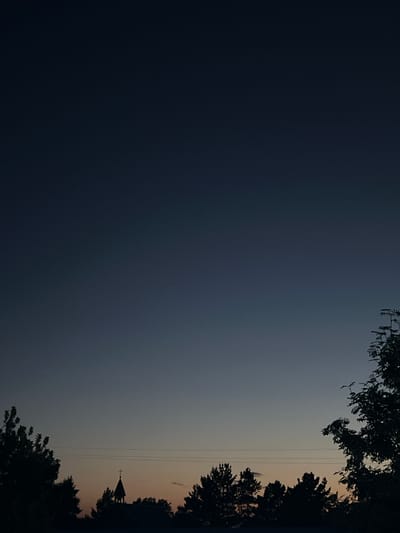 The image depicts a twilight sky over Sherbakul, Russia. Silhouetted against the gradient of dark blue to pale orange sky are several trees, a building topped with a cross, and power lines. The scene is calm and still, with no people visible. The focus is on the natural transition of the sky and the sparse, dark outlines of man-made structures and vegetation.
