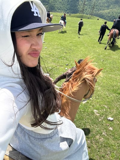 A selfie, slightly rotated, shows a woman wearing a dark blue baseball cap with a white logo, and a white hooded sweatshirt with grey sleeves. She is positioned in the foreground, with her face angled towards the camera. Behind her, partially blurred, is a brown horse with a bridle. In the background, several people are walking and riding horses on a green, grassy field. The landscape suggests a mountainous or hilly area. This scene is located in Teghut, Armenia.