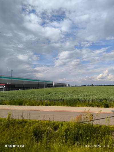 The image, taken in Robakowo, Poland on June 1, 2026 at 17:11, features a gray and green industrial building behind a green field. A gray road with a red-orange border runs in front of the field, followed by green foliage in the foreground. The sky is partially cloudy. The photograph was taken with a Xiaomi 13T.