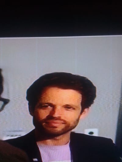 This image depicts a head-and-shoulders view of an adult male in a formal or semi-formal setting, located in Mista Ali, Nigeria. The subject, appearing to be Caucasian, has dark, wavy hair and a short beard or stubble. His forehead exhibits visible beads of moisture or sweat. He is wearing a dark-colored jacket over a light-colored, possibly lavender, t-shirt or collared shirt, with a small badge or pin visible on his left lapel. The man looks directly forward with a slight smile. The background consists of a plain light grey wall. Behind the subject's right shoulder, a white electrical outlet or switch plate with a dark circular element is mounted on the wall. The upper portion of the frame is black, indicating a screen edge or framing device. The image quality displays digital artifacts, pixelation, and scan lines, consistent with a photograph taken of a digital display.