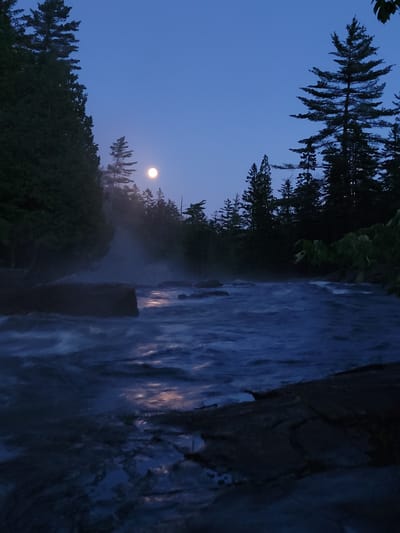 The image shows a river flowing through a forest at dusk or night, with a full moon visible in the sky. The water is dark blue and appears to be moving rapidly, with whitecaps and reflections of the moon. There are trees on both sides of the river, and some rocks are visible in the water. The sky is a deep blue, with a few wispy clouds. The overall mood of the image is peaceful and serene.
The location is likely in Saint-Alban, Canada, as indicated by the user. However, without more specific information or visual cues within the image itself, it is impossible to pinpoint the exact location within Saint-Alban. The scene is a natural environment, possibly a park or wilderness area, with a river and a forest.