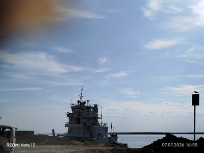 The image depicts a waterfront scene in Nizhny Bestyakh, Russia, on July 7, 2026, at 16:55. A large, multi-story white tugboat is moored near the shore. In the foreground, there is a pile of dark soil and rocks. To the left of the tugboat, the front of a white truck is visible, along with a dark, flat-topped structure. The water extends to the horizon, meeting a clear blue sky with scattered white clouds. A dark post with a bird perched on top stands to the right of the frame. The lower-left corner displays "REDMI Note 13" and the lower-right corner shows the date and time.