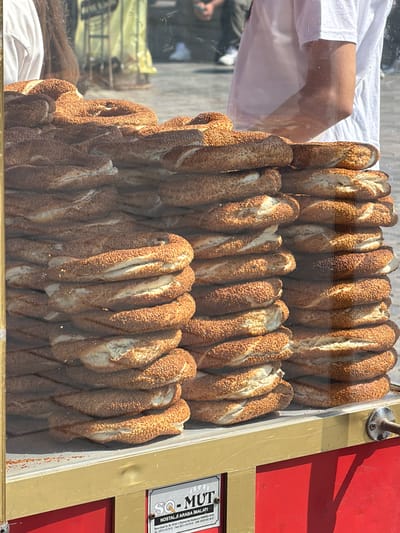 A display case filled with stacked simit, a Turkish bread, is positioned in front of a vendor selling them in the Fatih district of Istanbul, Turkey. The simit are round and covered in sesame seeds, with some appearing darker and crispier than others. Behind the display case, a person wearing a white t-shirt and jeans is visible, with their right arm slightly bent as if serving or preparing the simit. The background shows a blurred street scene with people and buildings. A sign on the front of the display case provides vendor information, including "SQ - MUT NOSTALJİ ARABA İMALATI" and contact details. The overall scene suggests a busy street food vendor.