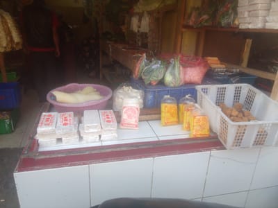 The image depicts a street-side market stall in Megamendung, Indonesia, likely a warung or small grocery store. In the foreground, a white tiled counter displays various food items. Stacks of wrapped white goods, possibly tempeh or tofu, are arranged in the lower left. To their right, several individually wrapped food items in pink and yellow packaging are visible. A pink basket containing light-colored, elongated objects, possibly more food items, is positioned behind the stacks of wrapped goods. Further to the right, a white plastic basket holds numerous small, brown, fried food items, likely fried tofu or tempeh. In the background, a person wearing a dark vest over a red shirt is partially visible, standing behind the counter. Shelves behind the counter are stocked with produce and other goods, including leafy green vegetables and what appear to be bags of snacks or other packaged items. The overall setting suggests a typical Indonesian market scene, with a focus on fresh and prepared food items for sale.