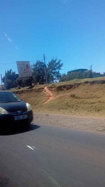 A car is seen on a tarmac road with a white arrow pointing in the opposite direction. On the road's side, there is a grassy, eroded bank. A medical center billboard is visible near trees. A worn, narrow path leads from the road up the eroded bank. The sky is clear and blue. Utility poles and wires are present in the background.