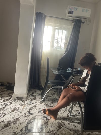 A young woman is seated at a black, glass-topped dining table in a room in Naze, Nigeria. She is wearing a white zip-up jacket over a dark top and dark shorts. The woman is looking down at a smartphone in her hands. The table has a black plate with some items on it and a plastic bottle. Two modern black chairs with metal legs are positioned around the table. Behind the table, sheer curtains are drawn across a window, with dark curtains layered in front of them. An air conditioning unit is mounted high on the wall above the window. The floor is tiled with a white and gray marbled pattern. The room appears to be a dining area.