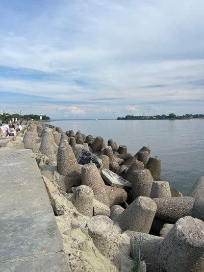 This image depicts a scene in Baltiysk, Russia, featuring a concrete breakwater composed of numerous tetrapods. The breakwater lines the edge of a body of calm water, likely the Baltic Sea. On the left side of the image, a concrete walkway runs parallel to the breakwater, with several people visible walking and sitting on it. Beyond the walkway and breakwater, a coastline with trees and buildings can be seen in the distance, including a prominent lighthouse. The sky is partly cloudy, with a mix of blue sky and white clouds. The overall impression is of a coastal recreational area, possibly a promenade or pier, designed to protect the shore from waves.