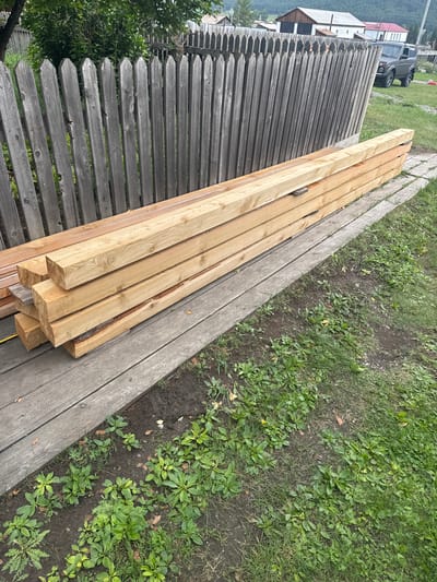 A stack of lumber lies on a wooden walkway next to a weathered wooden fence in Yabogan, Russia. The lumber, comprised of at least nine rectangular beams, is light brown and appears to be new. Behind the fence, trees and buildings can be seen. To the right of the walkway, grass and dirt are visible. A grey SUV is parked on a gravel area to the right of the fence, with buildings in the background.