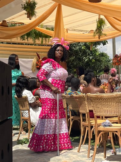 This image captures an outdoor social gathering in Kumasi, Ghana, under bright daylight conditions. The central subject is an older, dark-skinned woman with dark, curly hair, standing and facing towards the right side of the frame. She wears a floor-length gown of pink and white lace featuring geometric and floral patterns, an asymmetric neckline, and a short sleeve on her visible right arm. A pink fascinator hat with looped elements rests on her head. She holds a silver walking cane vertically in her right hand.
To the left of the central figure, a woman in a light blue patterned dress stands, smiling slightly. Further back and to the right, numerous other individuals, predominantly women, are seated or standing. Many are positioned in wicker-backed wooden chairs around tables that feature floral arrangements in shades of orange and pink. The event is hosted beneath a light orange fabric canopy, from which clusters of green foliage are suspended as decoration. A large, dark, rectangular speaker enclosure is visible on the far left of the foreground. The ground beneath the canopy is a paved surface. Lush green trees are visible in the distant background, beyond the event structure.