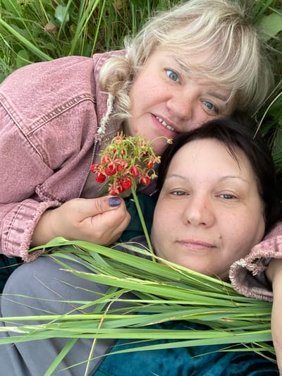 The image shows two women lying down in tall green grass. The woman in the foreground has dark hair and is wearing a dark green top and a pink jacket. The woman behind her has blonde hair and is wearing a pink jacket. She is holding a small cluster of red berries in her hand. The background consists of grass and some foliage. The scene appears to be outdoors, possibly in a rural or natural setting near Vyškov, Russia.