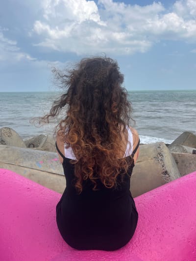 A woman with long, curly brown hair is seated facing away from the camera, looking out at a choppy grey sea under a cloudy sky. She is wearing a black dress with a white strap visible. The woman is positioned on a bright pink, curved structure, which appears to be part of a public seating area or art installation. Behind her, grey concrete tetrapods line the shore, serving as a barrier against the waves. The sea is a deep grey, with white foam crashing against the tetrapods. The sky is filled with dynamic, textured clouds in shades of white and grey, with patches of lighter blue peeking through. The overall impression is one of a person contemplating the vastness of the ocean. This scene is not located in the city of Phú Thủy, Vietnam.