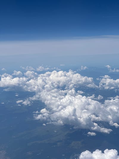 An aerial view captures a vast expanse of the sky, transitioning from a deep, vibrant blue at the top to a lighter, hazy blue near the horizon. Below the sky, a dense layer of fluffy white cumulus clouds dominates the frame, casting shadows on the terrain below. Peeking through the clouds, a mountainous landscape is visible, with patches of snow adorning the higher elevations. The topography suggests a rural or wilderness area, with subtle hints of human presence in the form of scattered structures and winding pathways that are faintly discernible through the haze and cloud cover. The scene conveys a sense of altitude and perspective, characteristic of an airplane's vantage point. There is no visual information to confirm or deny the specific location within Firajë, Kosovo.