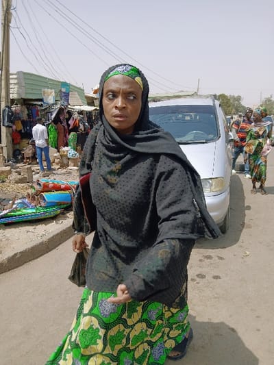 This image depicts a daytime street scene in the city of Kungulu School, Zambia. The primary subject, an adult female, is captured walking towards the viewer's right, her body angled in motion. She wears a black headscarf over a green and yellow patterned headwrap, a black textured long-sleeved top with a sheer black overlay, and a long green and yellow patterned skirt. Dark sandals are visible on her feet. Her eyes are wide and directed to her left, displaying a focused or slightly apprehensive expression. She holds a dark bag in her right hand; her left hand is partially open.
To the left of the frame, a series of market stalls line a dirt and paved area adjacent to the street. These stalls display various clothes and rolled fabrics. A light green corrugated metal structure is visible among them. A utility pole with multiple overhead wires stands prominently on the far left. A male figure in a white shirt and blue jeans walks past these stalls. In the mid-ground, a silver minivan is positioned on the dusty street. Further behind the vehicle, several pedestrians, including two women in patterned dresses and a man in a striped shirt, are visible. Shadows on the unpaved ground indicate a sunny, possibly hazy day.
