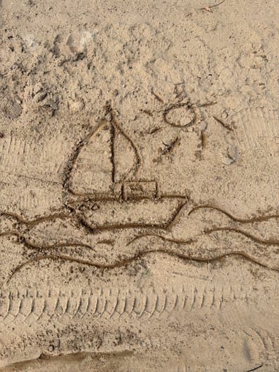 The image depicts a sandy surface with drawings etched into it. In the upper right quadrant, a rudimentary sun is drawn with rays emanating from a circular shape. To the left and slightly below the sun, a sailboat is depicted. The sailboat has a hull, a mast, and a sail. Below the sailboat, wavy lines represent water. Towards the bottom of the image, tire tracks are visible in the sand, suggesting that a vehicle has passed through this area. The overall scene is outdoors, on a beach or sandy terrain. The location is identified as Upeslejas, Latvia.