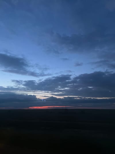 The image shows a landscape at dusk with a dark foreground of fields and a horizon line. Above the horizon, there is a strip of red-orange light, indicating the setting sun. The sky is a deep blue, with scattered dark clouds. There are a couple of faint vertical structures visible on the horizon line, possibly power poles or communication towers. The scene is set in the city of Belkopа, Kazakhstan.