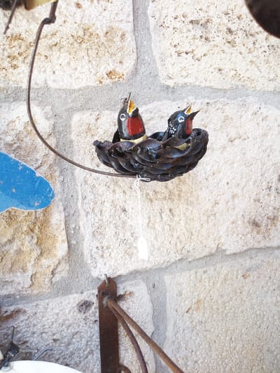 The image depicts a close-up, vertical shot of decorative metal birds in a nest, set against a stone wall. The nest is made of dark, possibly rusted metal, with a textured, layered appearance. Two small, colorful metal birds are perched inside the nest. One bird, on the left, has its beak open, revealing a yellow interior and red markings on its chest. The other bird, to the right, also has its beak open and appears to have black and white speckled detailing on its head. A thin white string hangs down from the nest, with a small white tag attached to its end. The background is a rough, textured stone wall, composed of irregular, light-colored blocks with visible mortar lines and patches of discoloration. A thin, dark metal wire curves from the upper left of the frame towards the nest, suggesting it's part of a larger hanging decoration. To the left of the nest, a flat, blue, wing-shaped metal piece is partially visible. A rusted metal bracket is attached to the stone wall in the lower right of the frame, from which another curved metal element extends. This scene is likely located outdoors, possibly as a decorative element on a building or in a garden, in the historic town of Besalú, Spain, known for its well-preserved medieval architecture.