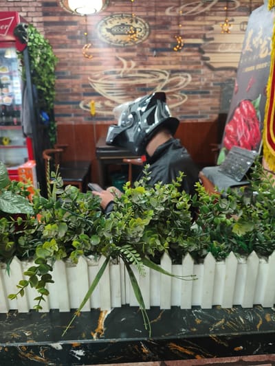 An individual wearing a black motorcycle helmet and a black jacket is seated at a table, looking down at a smartphone. They are positioned behind a decorative white picket fence with artificial green foliage. In the background, a brick wall is adorned with graphics of a steaming cup of coffee, a crescent moon, and stars. To the right of the individual, a black tablet is propped up on a grey surface. A red image of what appears to be crayfish is visible on a poster in the far right of the frame. The scene takes place in a dimly lit indoor setting that resembles a restaurant or cafe, possibly located in Jixi, China.