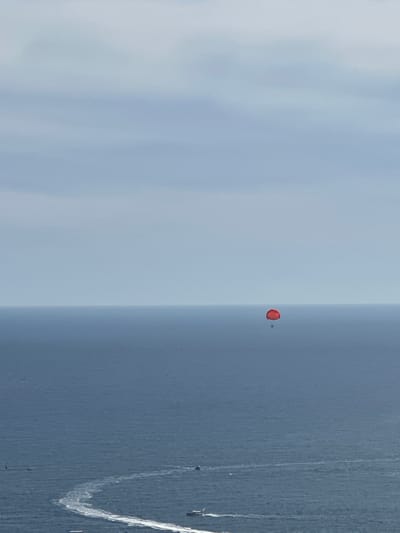 A parasailer in a red canopy is suspended over the vast, deep blue Adriatic Sea, off the coast of Vrela, Montenegro. The parasailer is a small, solitary figure against the immense backdrop of the ocean and the pale, cloudy sky, which occupies the upper two-thirds of the frame. The horizon line is sharp, dividing the water and sky. Closer to the viewer, the water is disturbed by the wakes of several small boats, leaving trails of white foam on the surface. The scene conveys a sense of aerial freedom and the expansive natural beauty of the Montenegrin coastline.