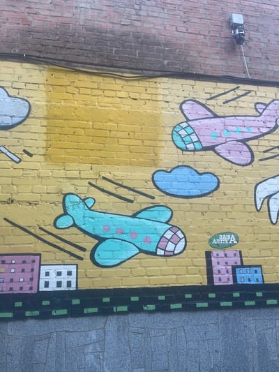 The image displays a section of a yellow brick wall adorned with graffiti. The graffiti depicts cartoonish airplanes in flight, clouds, and stylized buildings. One airplane is pink with teal circles, and another is teal with pink circles. Both airplanes have checkered wing tips. Several black lines suggest the movement of the planes. Blue clouds are also visible. Below the aerial scene, the graffiti continues with representations of buildings with pink and white facades, and a row of black and green rectangular shapes. To the right of the central area, a green oval-shaped sign with white lettering reads "ВАШ АПТЕКА," which translates to "YOUR PHARMACY." The wall is situated in the village of Horozhuli, Ukraine. The brick wall is partially visible at the top, with a darker colored brick pattern. A security camera is mounted on the upper section of the brick wall.