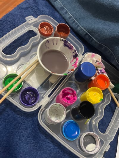 The image shows a close-up view of a clear plastic paint case filled with various colors of paint, brushes, and a crumpled piece of paper with paint splatters. The paint case is open and resting on a textured blue surface, likely a blanket or cloth. Inside the case, there are small containers of paint, some with lids on and some open. Colors visible include green, purple, pink, yellow, orange, blue, and black. There is also a larger cup or bowl containing a greyish-purple liquid, with streaks of purple and pink paint on its rim. Two wooden paintbrushes are positioned across the green and purple paint containers. A crumpled piece of white paper with colorful paint splotches is visible near the orange and blue paint containers. The background shows a section of blue denim fabric. The overall scene depicts an art or craft activity in progress. The image does not provide any information about the location in San Luis, Peru.