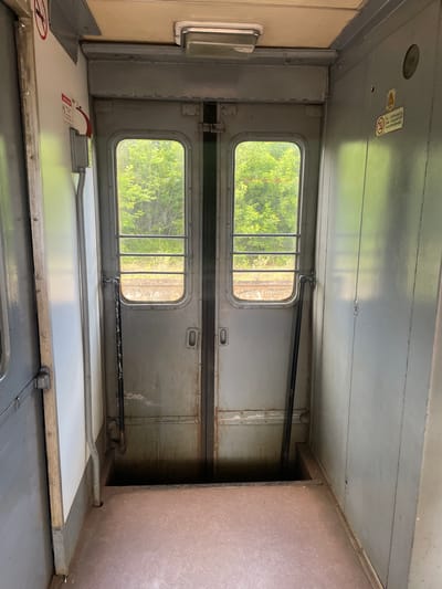 This is a vertical, eye-level, full shot of the interior of a train car in Блужскі сельскі Савет, Belarus. The image focuses on a pair of double doors with windows that lead to the exterior of the train. The doors are made of a weathered, gray metal and have horizontal bars across the windows for security. Through the windows, lush green trees and foliage are visible, indicating the train is in a rural or wooded area. The floor inside the train car is a worn, reddish-brown material with a speckled texture. There is a dark opening beneath the doors, suggesting a gap between the train car and the platform or ground. The walls of the train car are also gray metal, showing signs of wear and tear. On the left wall, there's a white panel with some red signage and a fire alarm. On the right wall, there's a warning sticker with Cyrillic text and a symbol of a person falling. The lighting appears to be natural light coming from the windows, supplemented by an overhead fluorescent light fixture. There are no people visible in the image.