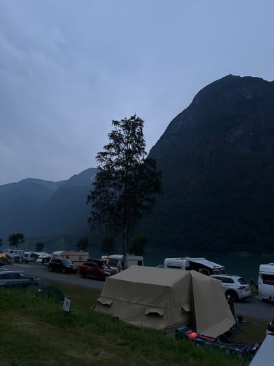 In the city of Olden, Norway, a campsite is situated at the base of a massive, dark, forested mountain that rises steeply from a calm, teal body of water. The sky is a hazy, overcast gray, suggesting early morning or late evening. A tall, slender tree with sparse leaves stands prominently in the foreground, silhouetted against the sky and mountains. The campsite is populated with several recreational vehicles, including caravans and campers, and a few cars. A large, tan canvas tent is set up in the foreground, with some clothing and camping gear laid out nearby. The overall impression is one of a tranquil, natural setting with human presence through the campsite.