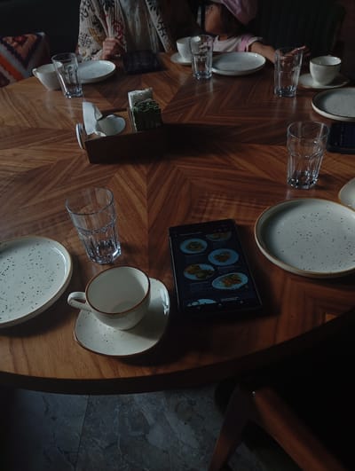 The image shows a close-up view of a round wooden table set for a meal in what appears to be a restaurant. The table has a distinctive wood grain pattern. On the table are several white ceramic plates with speckled brown details, and matching coffee cups and saucers. There are also several clear glasses, some filled with water, suggesting that drinks are being served. A phone is placed on the table, displaying an image of food dishes, likely a menu. In the background, two people are partially visible, suggesting a dining activity. One person appears to be an adult wearing a patterned top, and another is a child wearing a pink hat. The lighting suggests it might be daytime, possibly late afternoon or early evening, with some areas of the scene in shadow. The overall impression is a casual dining experience. The visible text on the phone screen is in Cyrillic script, with words like "Жар" and "Гранд" discernible, potentially indicating dishes or a restaurant name. The context provided mentions Бактуу-Долоноту, Kyrgyzstan, which places the scene in that specific geographical location.