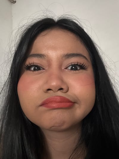 A close-up, frontal, waist-up shot shows a young Indonesian woman with long, dark hair and fair skin. She is wearing heavy eye makeup, including false eyelashes, and coral-toned lipstick. Her cheeks are heavily rouged with blush. Her expression is one of slight pout or displeasure, with her mouth downturned. Her eyes are wide open and looking directly at the camera. The background is a plain, white wall. The image was taken in North Jakarta, Indonesia.