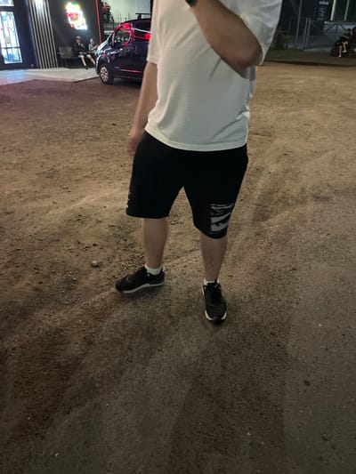 The image shows a person from the waist down, wearing a white t-shirt, black shorts, and black athletic shoes with white accents. The person is standing on a gravelly surface. In the background, there is a dark-colored van and a building with neon signs. Further in the background, two individuals are seated on a bench next to the building, and another group of people are on a scooter. The scene appears to be outdoors at night, possibly in a commercial or entertainment area. The location is identified as Strumivka, Ukraine.