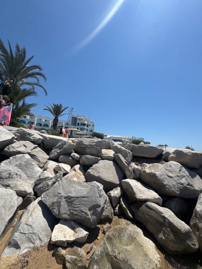 The image shows a bright, sunny day in Mrezga, Tunisia. In the foreground is a large pile of light grey rocks. Some rocks appear wet. To the left of the rocks, a clear blue sky is visible. In the mid-ground, behind the rocks, are white buildings and palm trees. Several people can be seen between the buildings and palm trees. A person holding a pink and black striped towel is visible in the lower left.