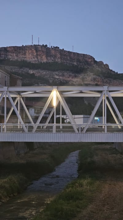 This image captures a view of a pedestrian bridge over a small river in Alhama de Aragón, Spain, during twilight. The bridge is constructed of white steel trusses and features a prominent vertical light fixture in its center. Beyond the bridge, a building with the illuminated "zalux" logo is visible, suggesting a commercial or industrial presence. In the background, a rugged, rocky hillside is topped with several telecommunications towers, indicating a more elevated or rural area surrounding the town. The river below is calm, with grassy banks on either side. The sky is a deep blue, characteristic of dusk. No people are present in the frame.